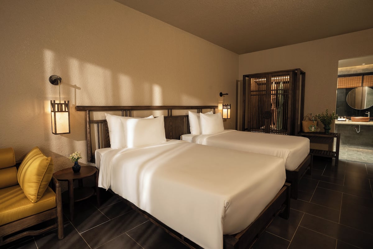 Habitación de hotel con dos camas, un sofá, una mesita con un florero, y un baño con espejo y lavabo.