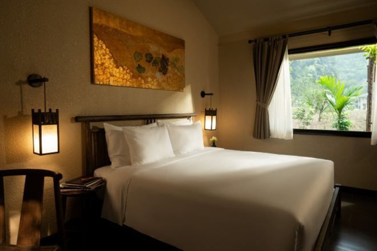 Een net opgemaakt bed in een hotelkamer, met een groot schilderij aan de muur, twee bedlampen en een raam met gordijnen.