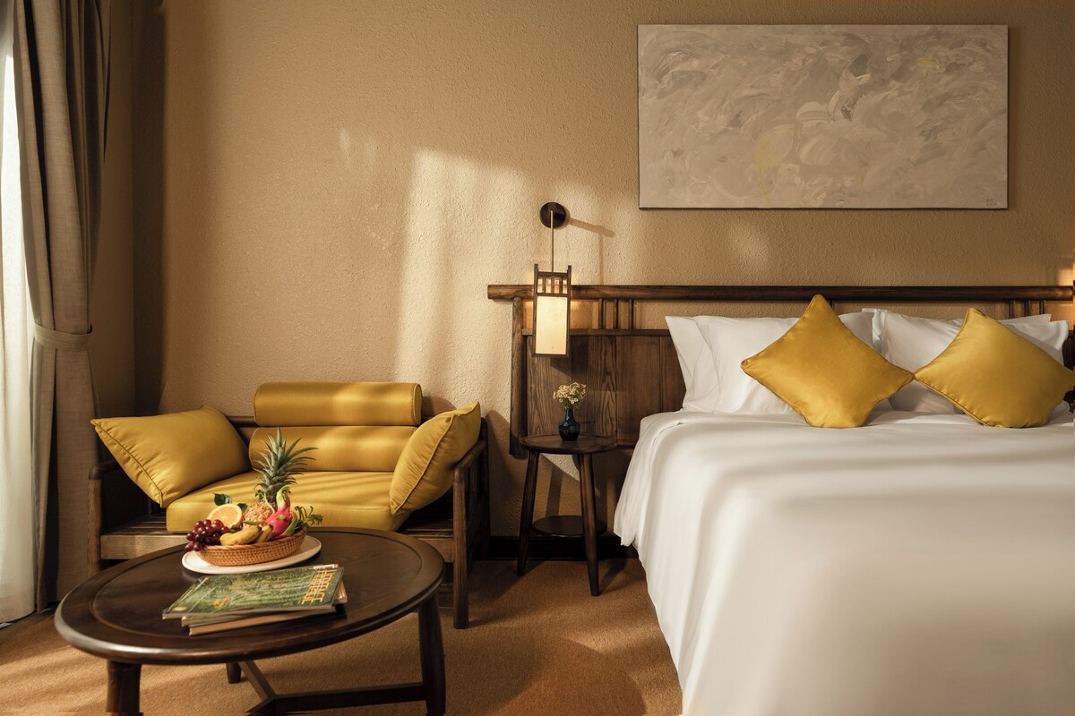 Une chambre d’hôtel avec un lit, un canapé, une table avec un panier de fruits et une petite table avec un vase.