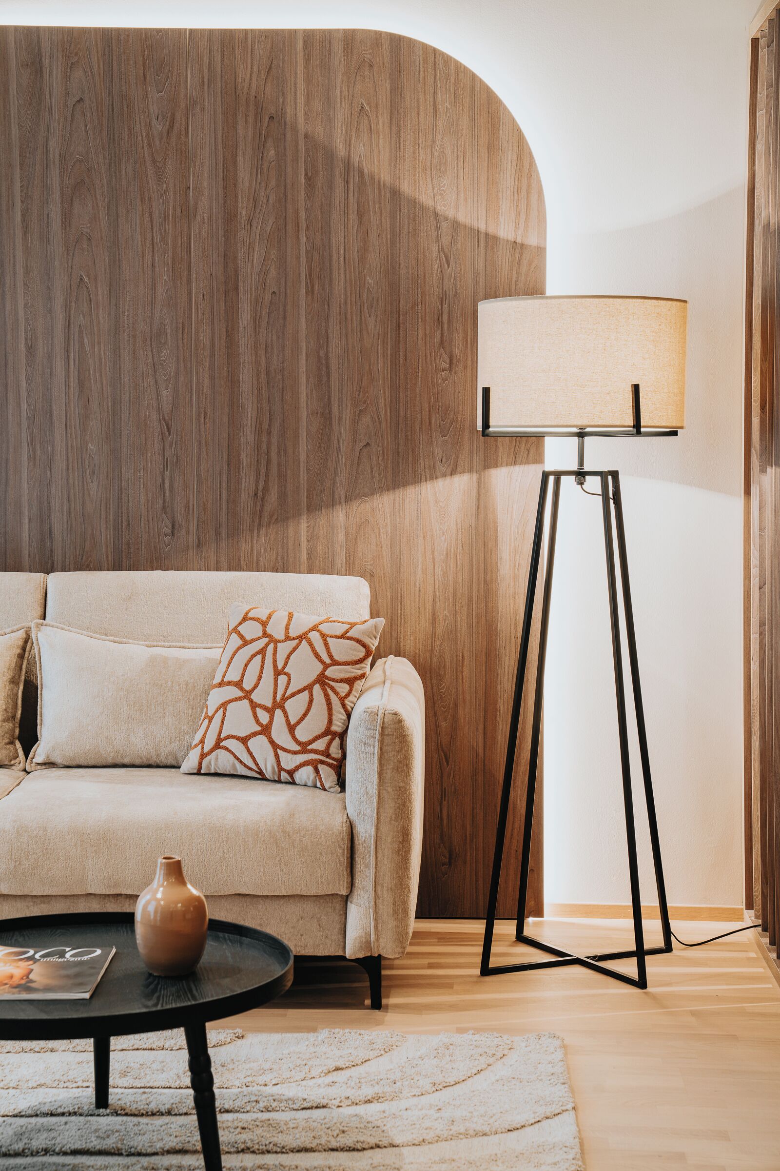 A modern living room with a wooden accent wall, a floor lamp, a sofa with patterned cushions, and a small table with a vase.