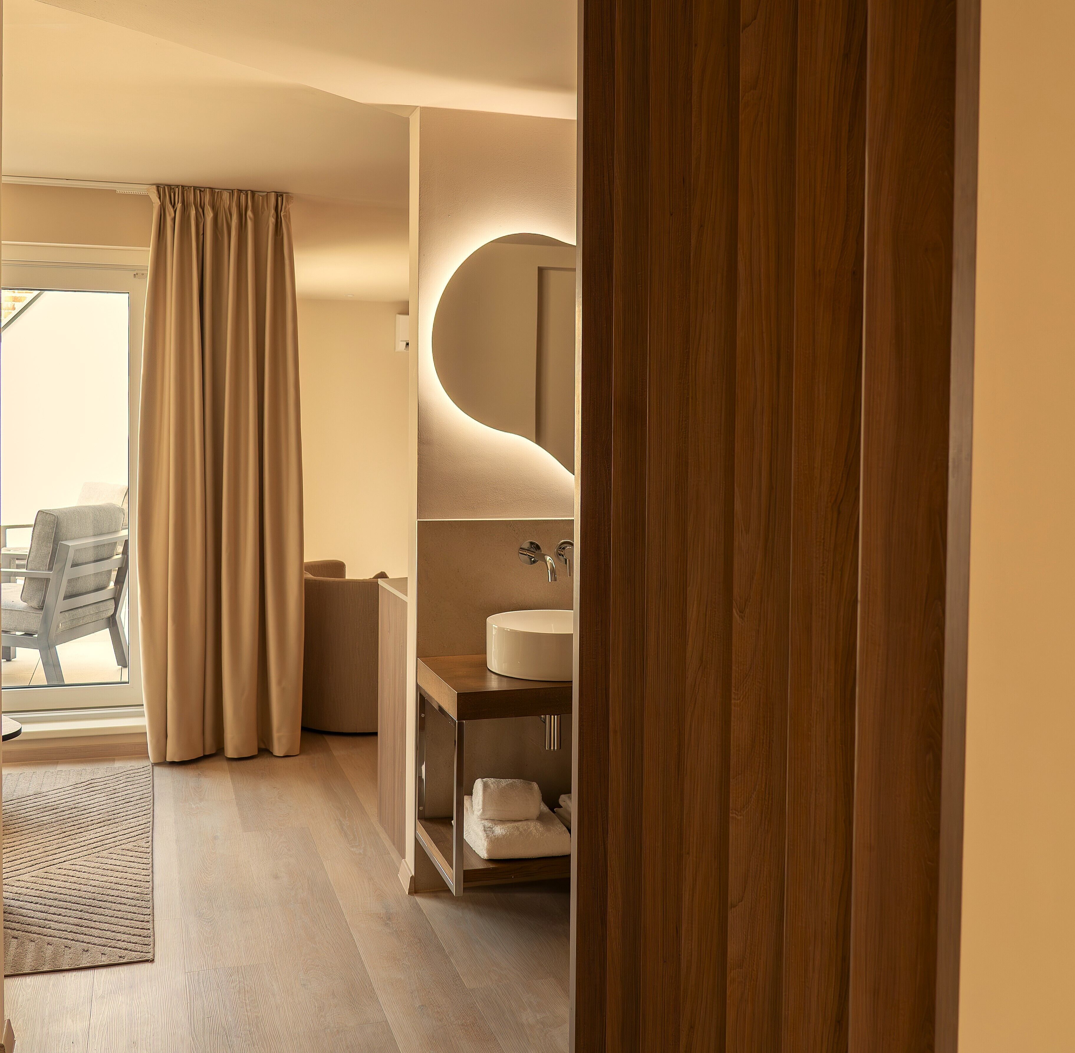 A modern bathroom with a wooden vanity, a round mirror, and a view of a balcony with a chair.