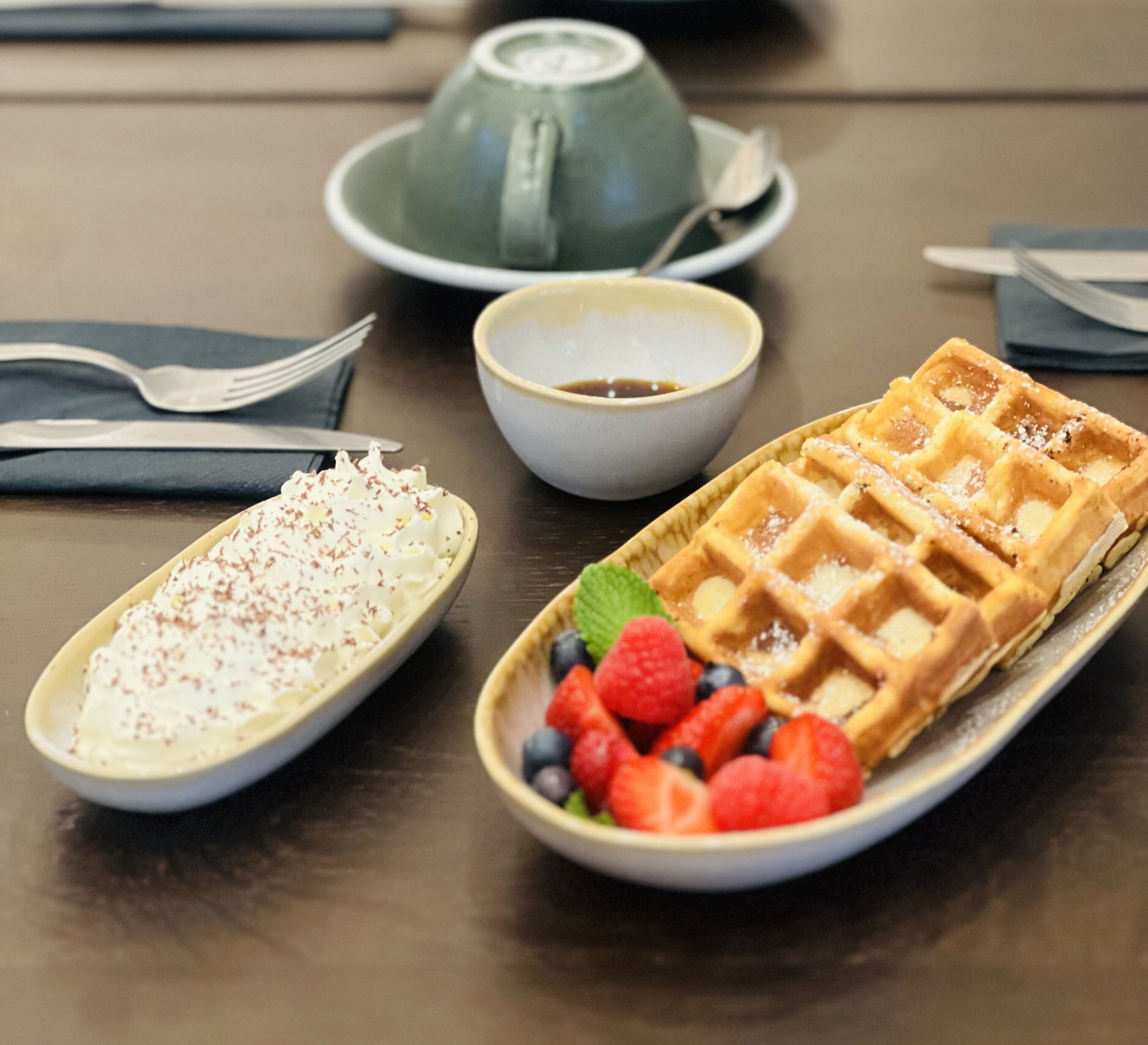 Una mesa con un gofre, un bol de fruta y un postre con crema batida.