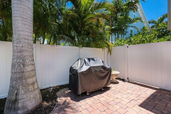 Ein Garten mit einem Grill, der mit einer Schutzhülle abgedeckt ist, einem weißen Zaun und einer Palme.