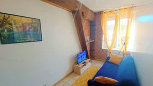 Une pièce avec un canapé bleu, un téléviseur posé sur un meuble en bois, un tableau accroché au mur et une fenêtre ornée de rideaux jaunes.