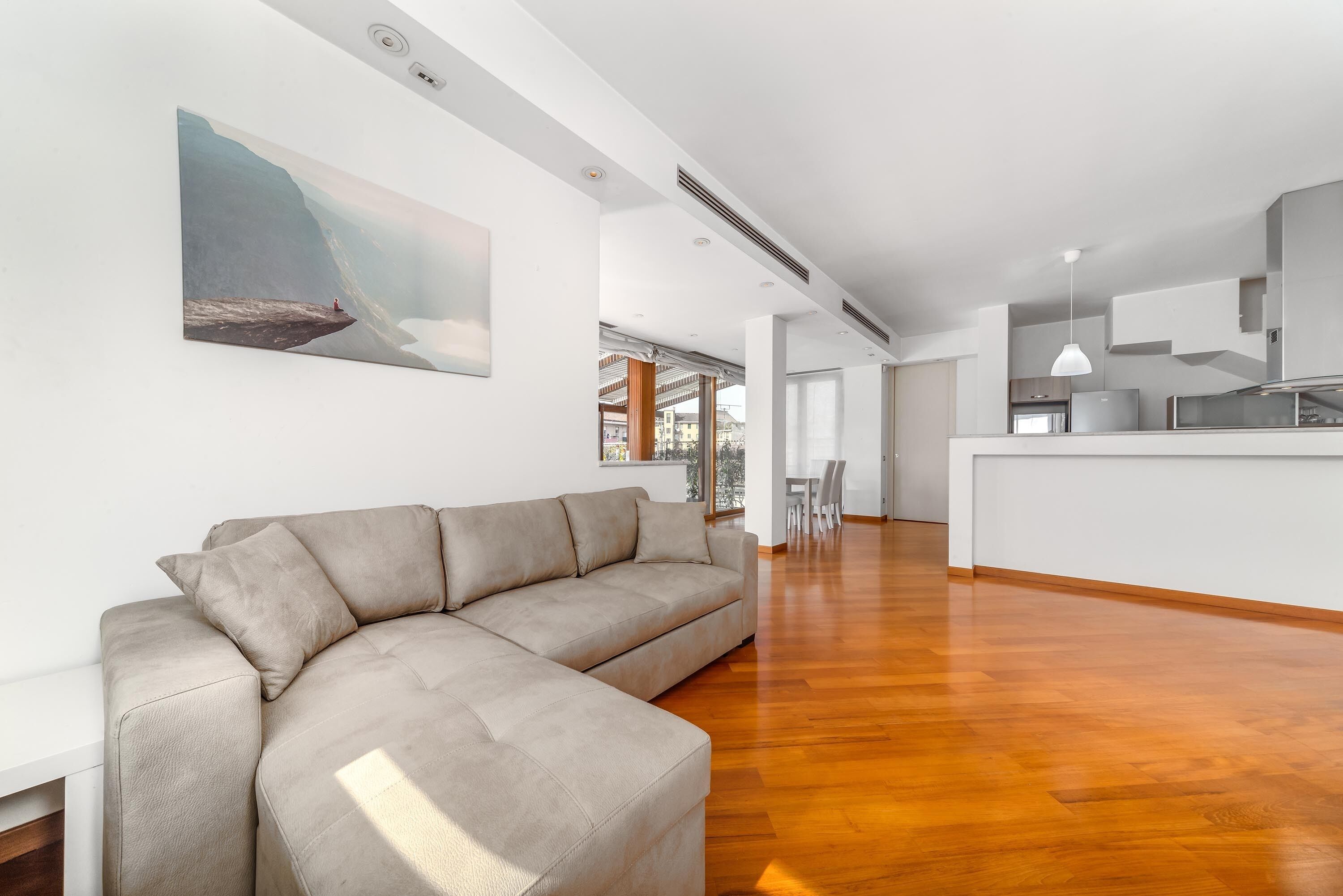A modern living room with a beige sectional sofa, a wooden floor, and a kitchen area with a white pendant light.