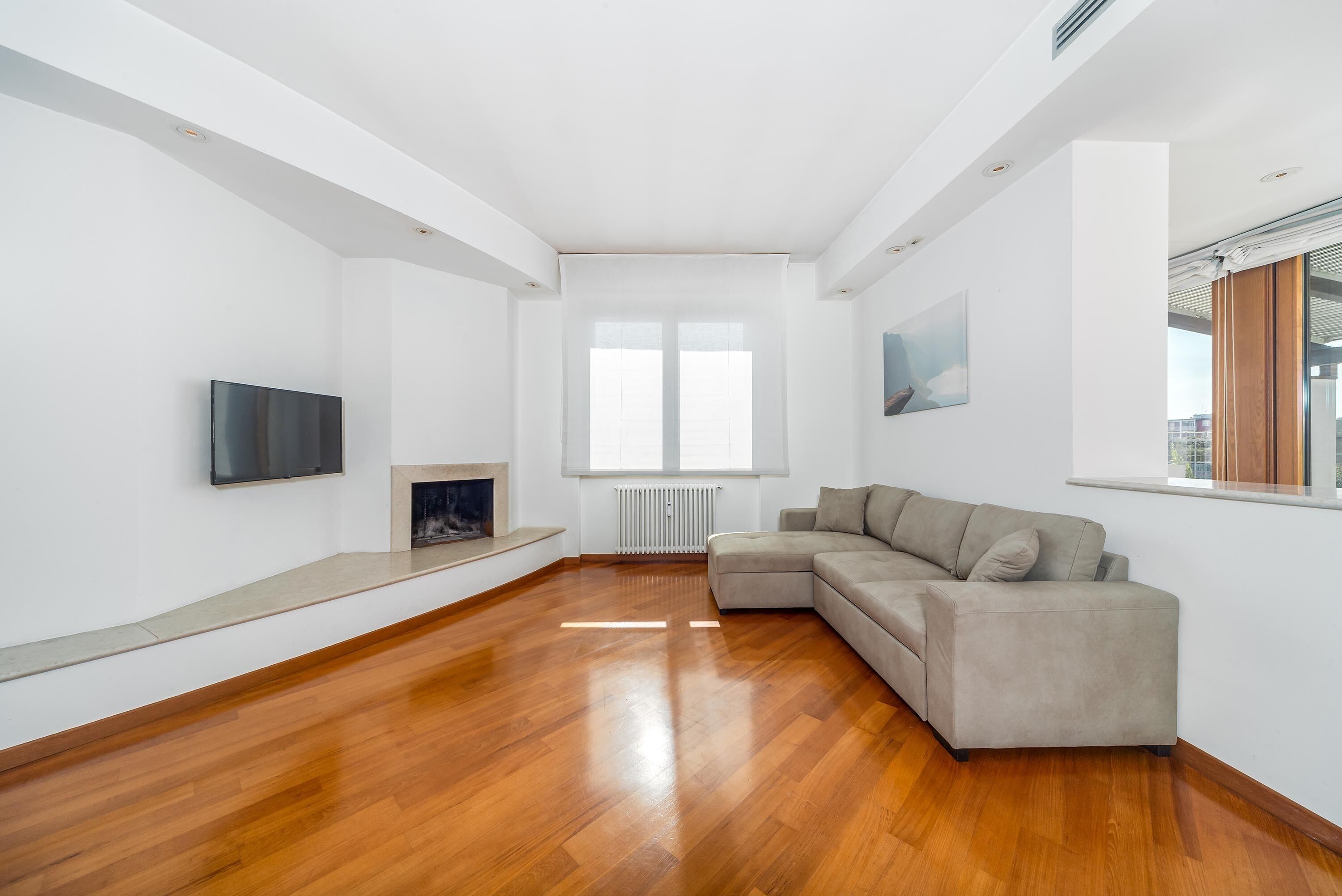 A modern living room with a wooden floor, a beige sectional sofa, a flat-screen TV mounted on the wall, and a fireplace.