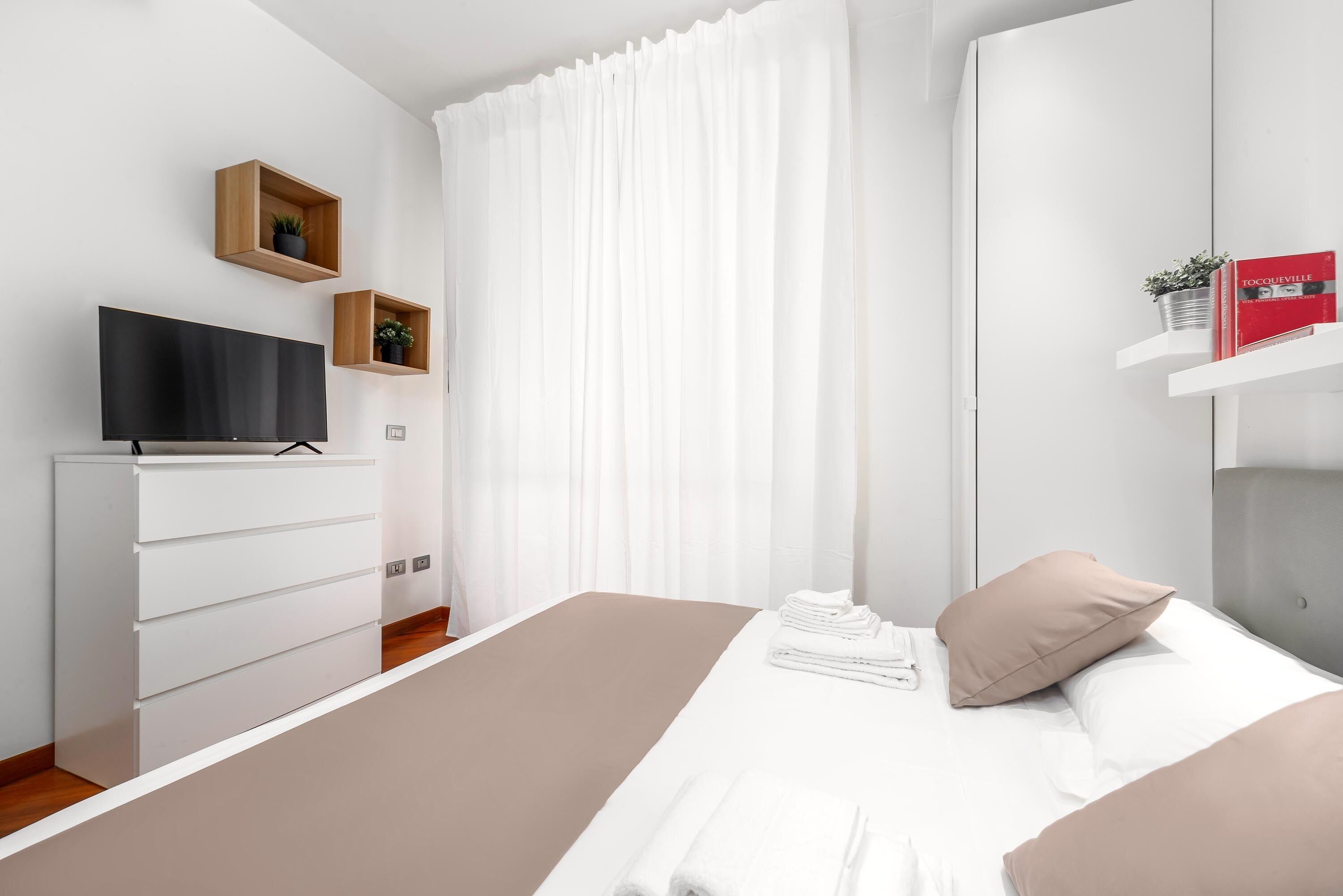 A modern hotel room with a flat-screen TV, a white dresser, a bed with white and beige bedding, and two wooden shelves with plants.