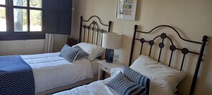 Ein Zimmer mit zwei Betten, jeweils mit weißer Bettwäsche und gestreifter Kopfkissen, einem Nachttisch mit Lampe sowie einem Fenster mit Vorhängen.
