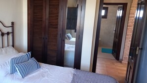 Ein ordentlich bezogenes Bett mit weißen und gestreiften Kissen, ein Holz-Kopfbrett und ein Blick ins Bad durch eine geöffnete Tür.