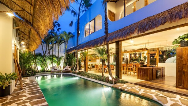 A swimming pool with stone tiles, surrounded by palm trees and a thatched roof structure.