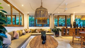 A modern living room with a wooden coffee table, a sofa with cushions, a hanging light fixture, and large windows with a view of greenery.