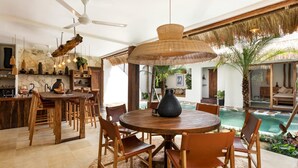 A spacious dining area with a round wooden table and chairs, a large woven pendant light, and a view of a pool outside.