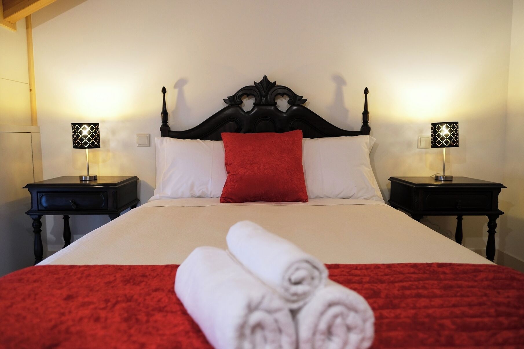 Een net opgemaakt bed met een rood-witte dekbedovertrek, twee nachtkastjes met lampen, en een opgerolde handdoek op het bed.