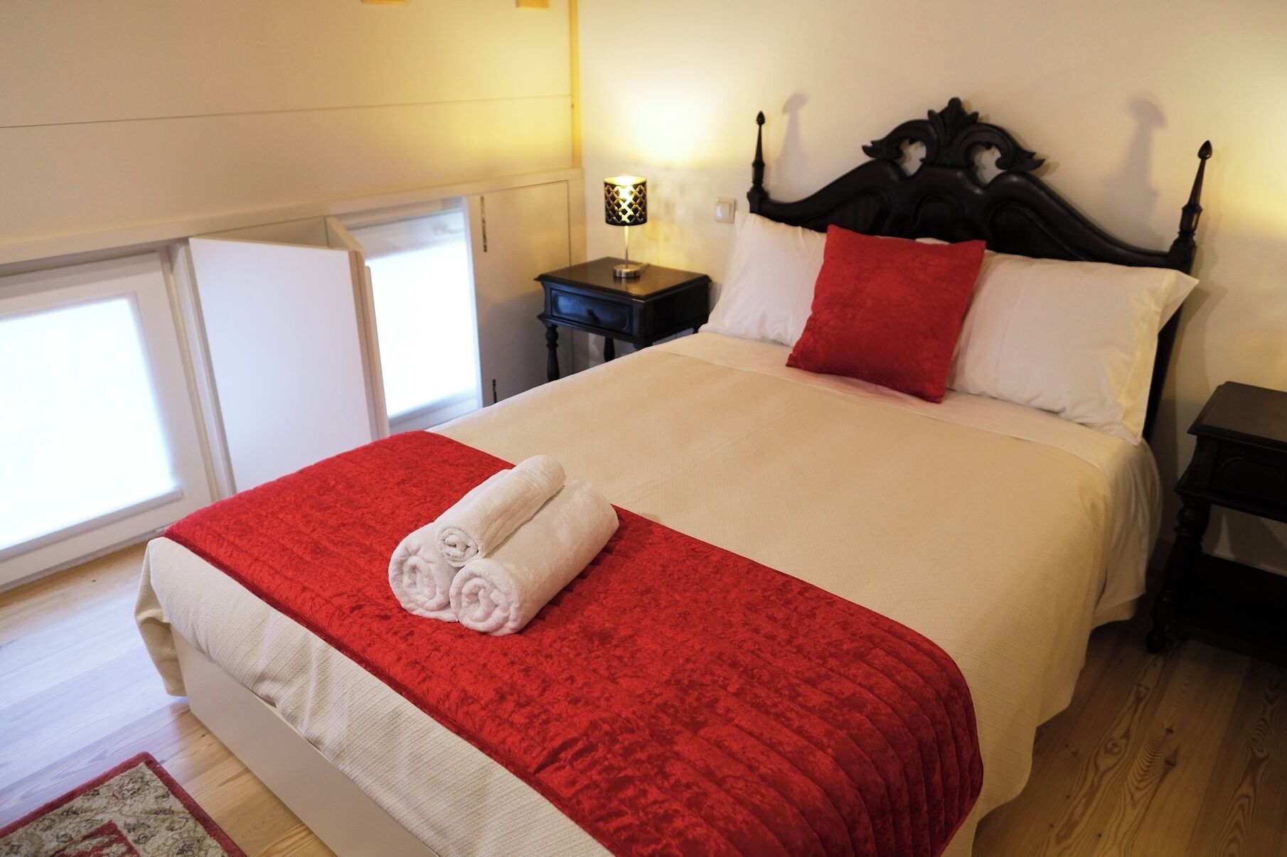 Un letto rifatto con cura, con copriletto rosso e bianco, una testiera nera e un comodino con una lampada.