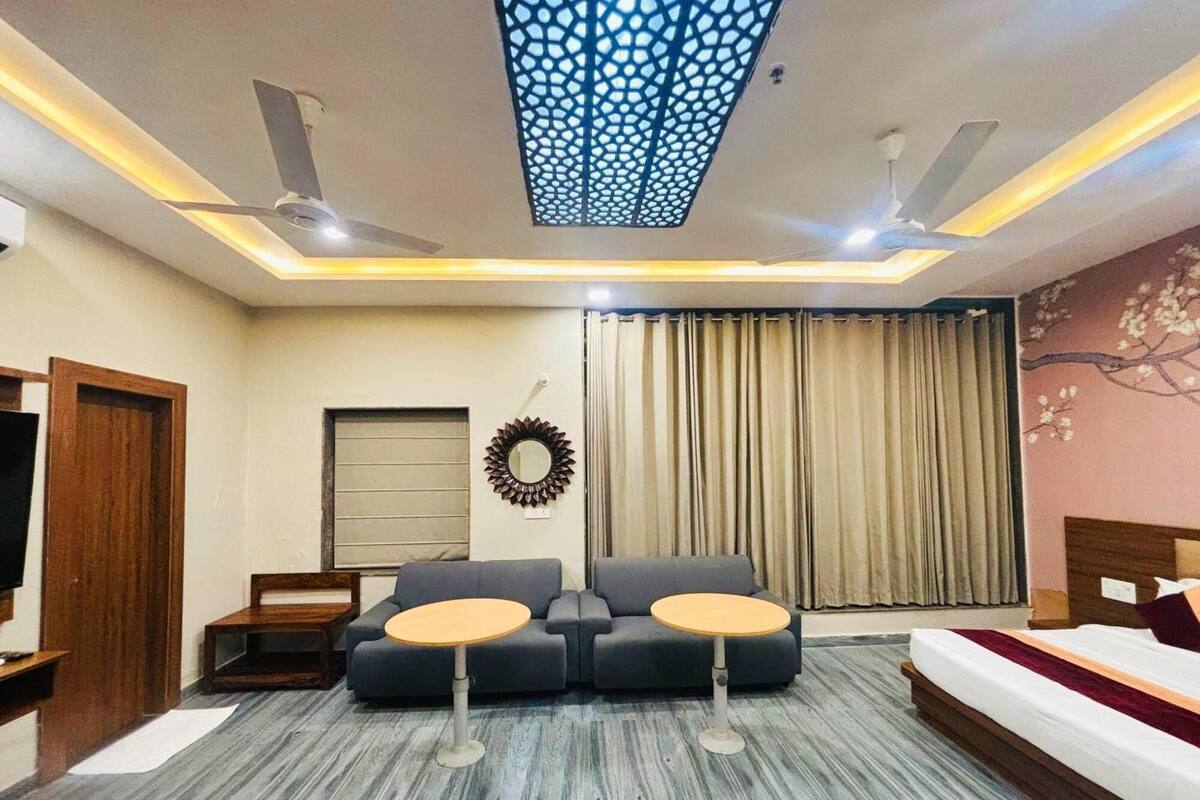 A modern hotel room with a sofa, two small tables, a bed, a TV, a ceiling fan, and a decorative wall piece.