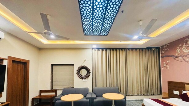 A modern hotel room with a sofa, two small tables, a bed, a TV, a ceiling fan, and a decorative wall piece.