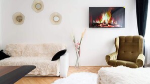 Un salon avec un canapé blanc, un fauteuil marron, une cheminée surmontée d’un tableau et un vase rempli de fleurs.