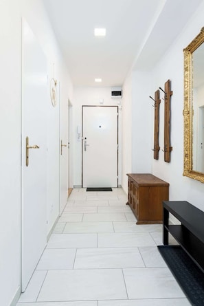 Un pasillo con piso de baldosas blancas, un banco de madera, un espejo grande con marco dorado y una puerta al final.