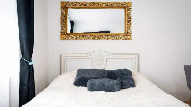 Ein Bett mit weißer, flauschiger Bettwäsche und zwei dunkelgrauen Kissen, darüber ein spiegel mit goldener Rahmung und ein weißer Kopfteil.