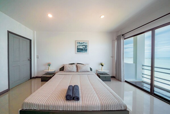 A modern hotel room with a large bed, a bedside table with a vase, a painting on the wall, and a balcony with a view.