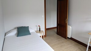 Et soveværelse med en seng, et lille natbord med en vase blomster, og en trædør der fører til et andet rum.