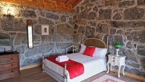 A bedroom with a stone wall, a wooden ceiling, a bed with white and red bedding, a nightstand with a lamp, and a dresser.
