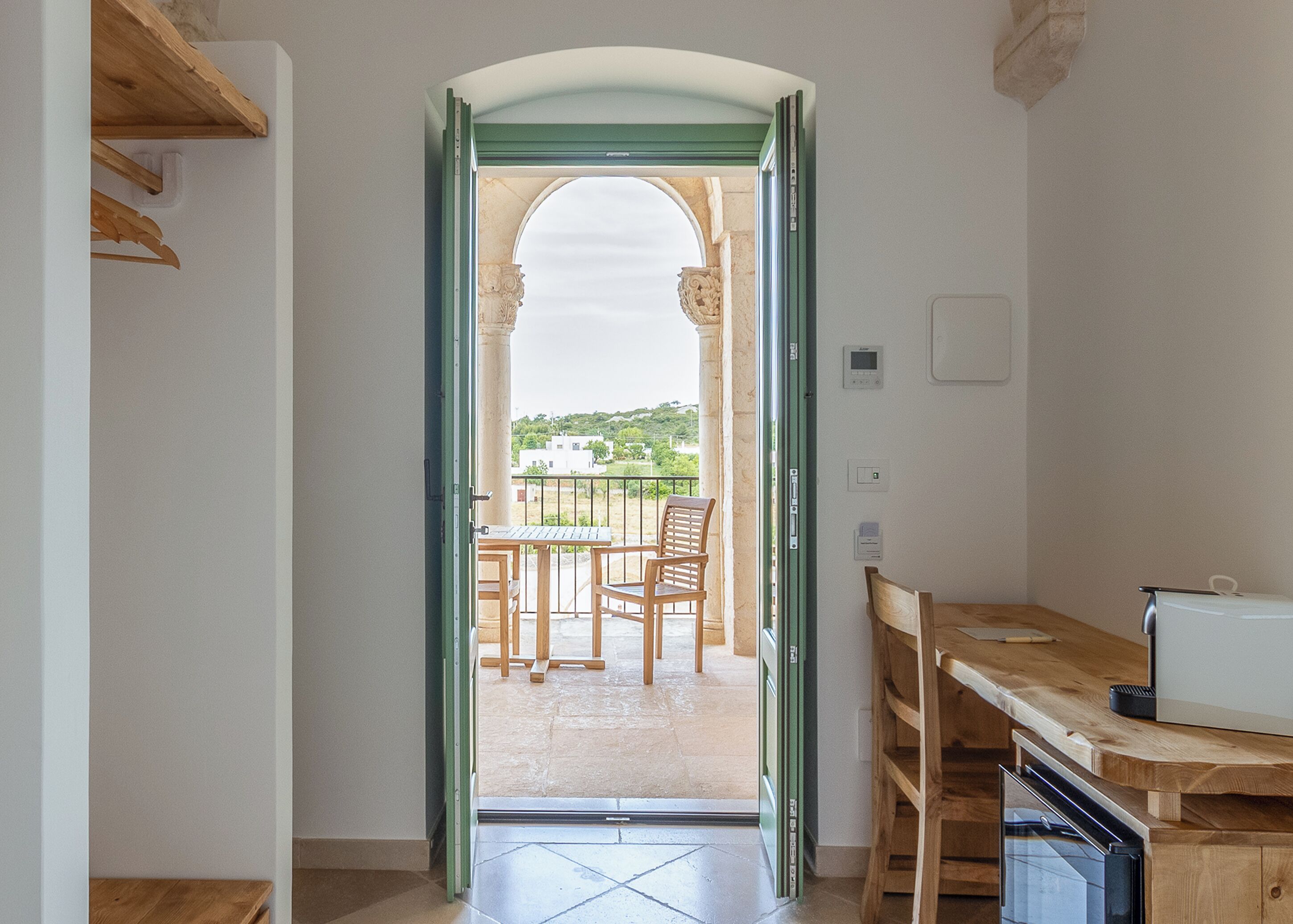 A narrow hallway leading to a balcony with a table and chairs, a wooden bench, and a view of greenery and buildings.
