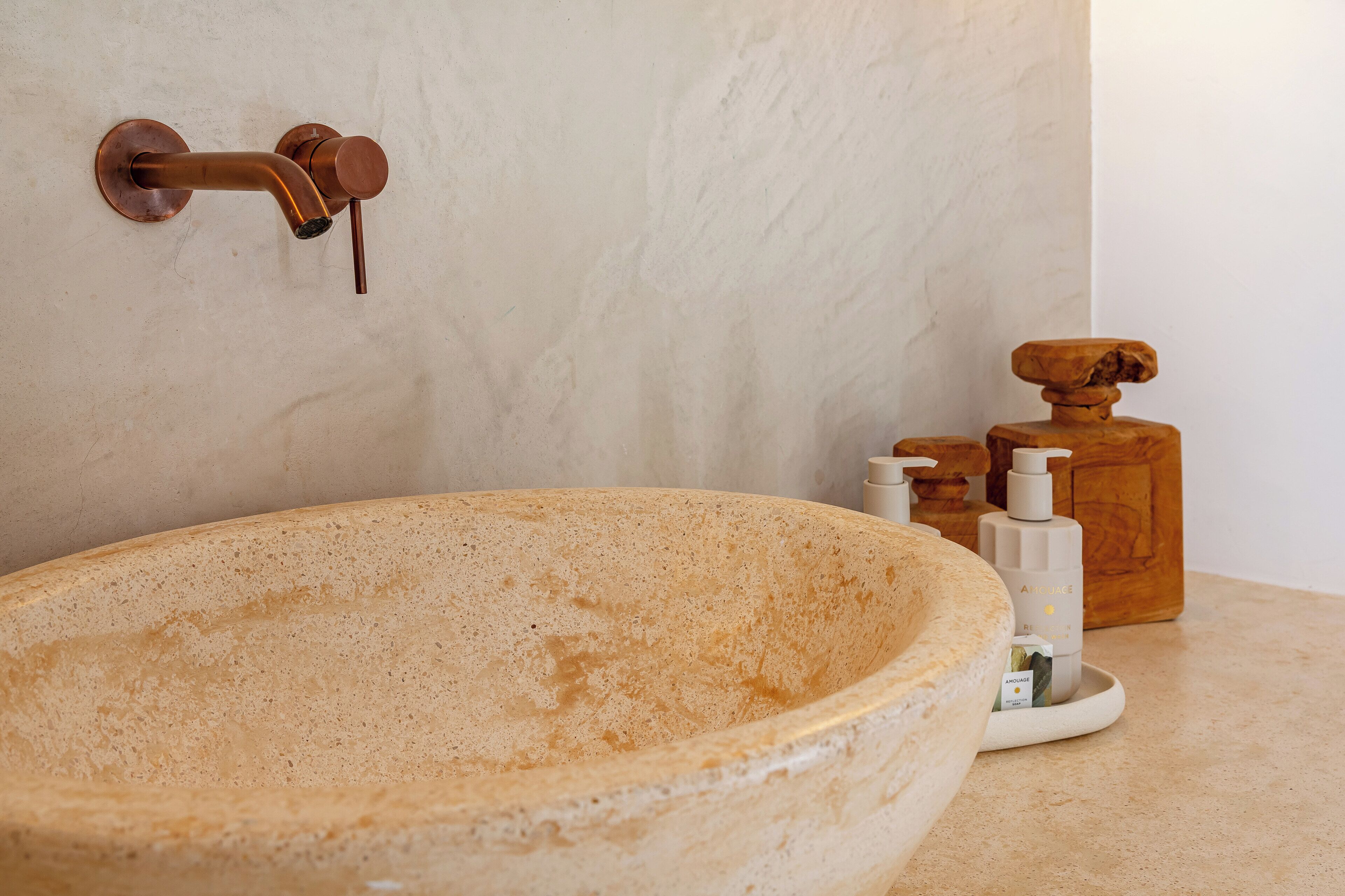 A bathroom with a large beige stone sink, a copper faucet, and a wooden soap dispenser on the countertop.