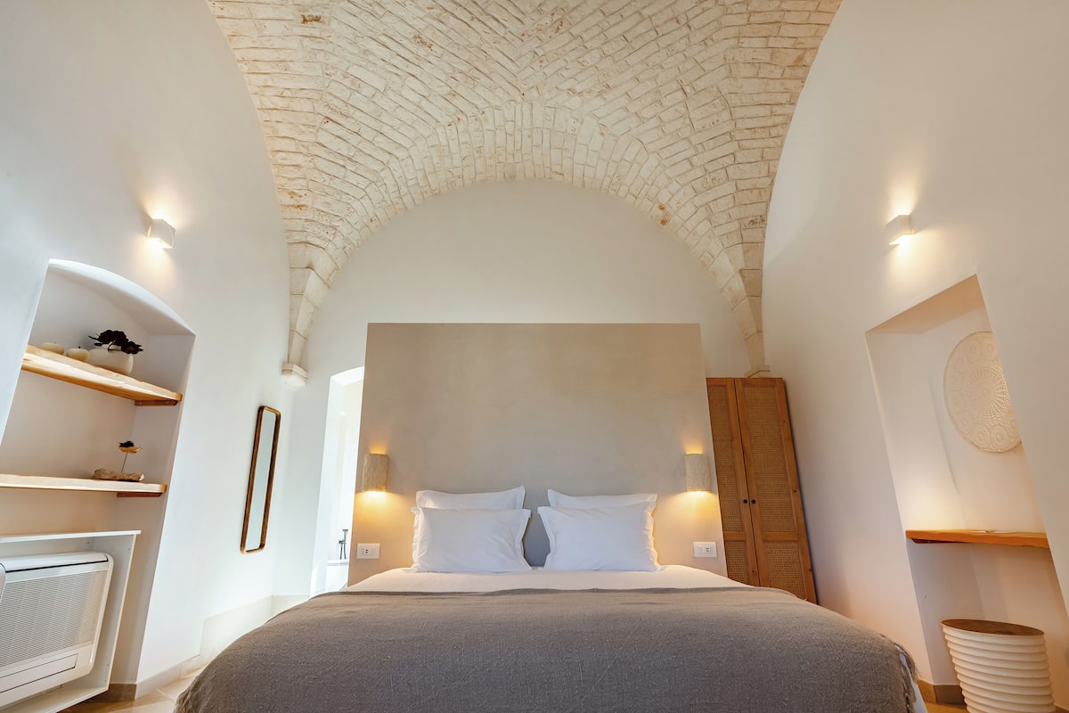 A bedroom with a brick ceiling, a bed with white bedding, a wooden wardrobe, and wall-mounted sconces.