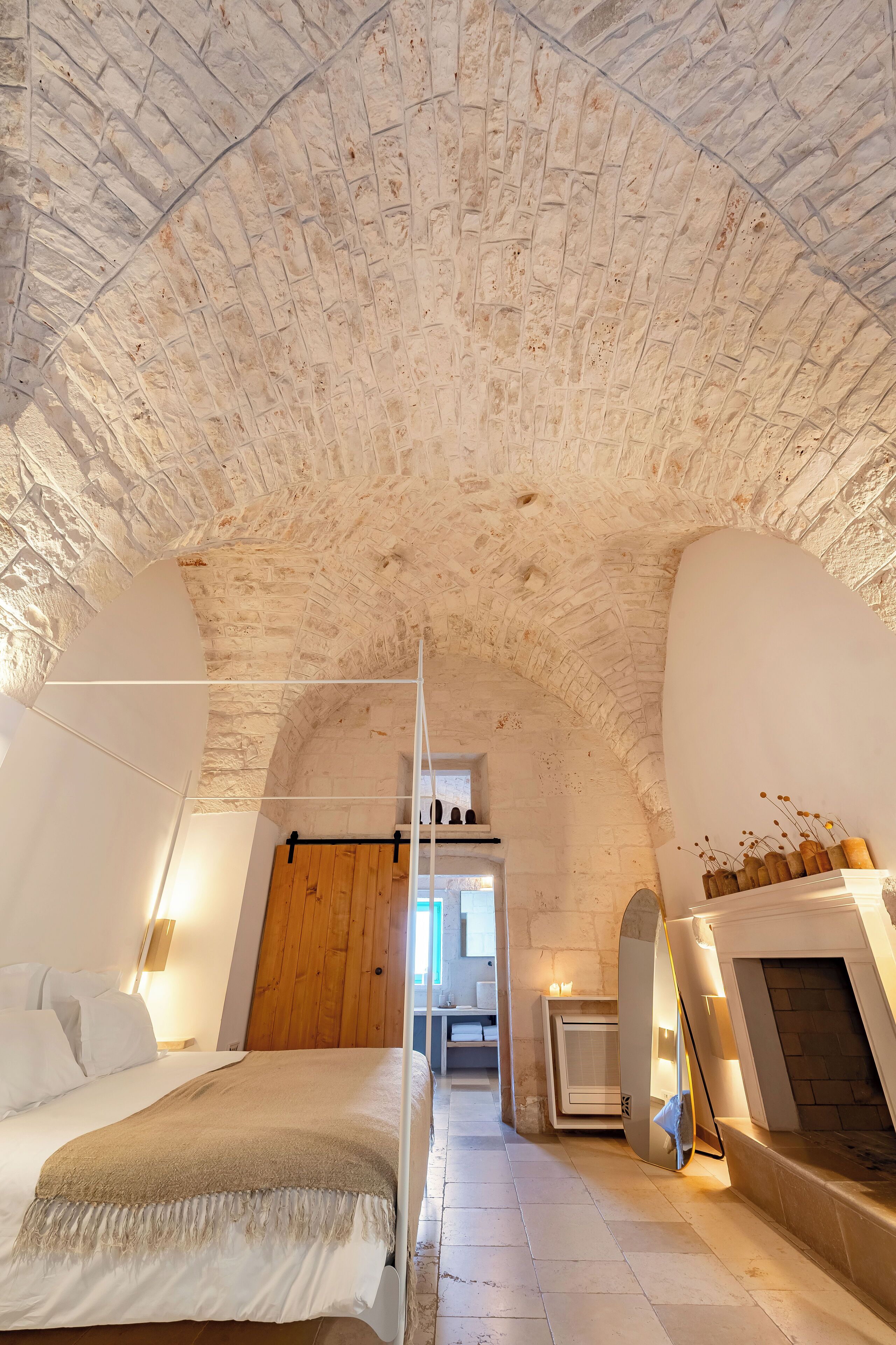 Ein Schlafzimmer mit einer gewölbten Decke, einem Bett mit einer beige farbenen Decke, einer Holztür und einem kleinen Tisch mit Kerzen.
