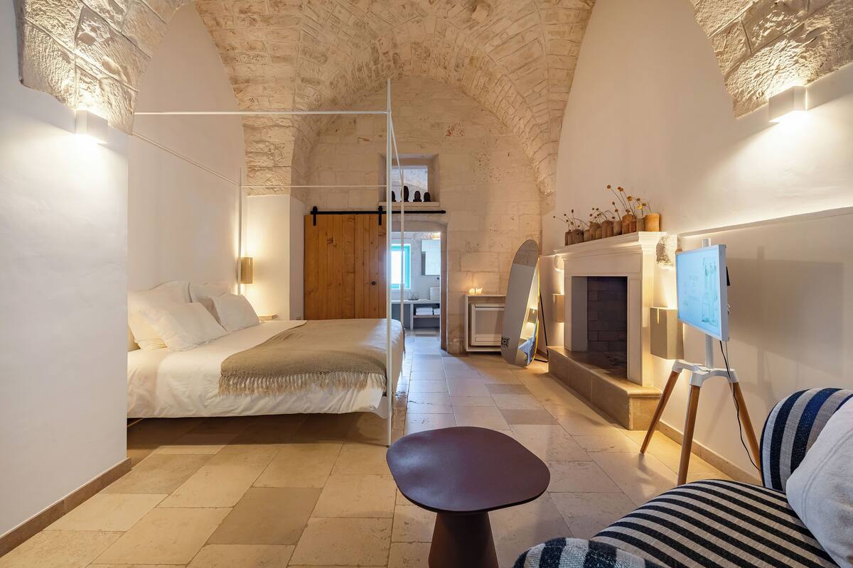 A bedroom with a bed, a small table, a sofa, a fireplace, and a painting on the wall.