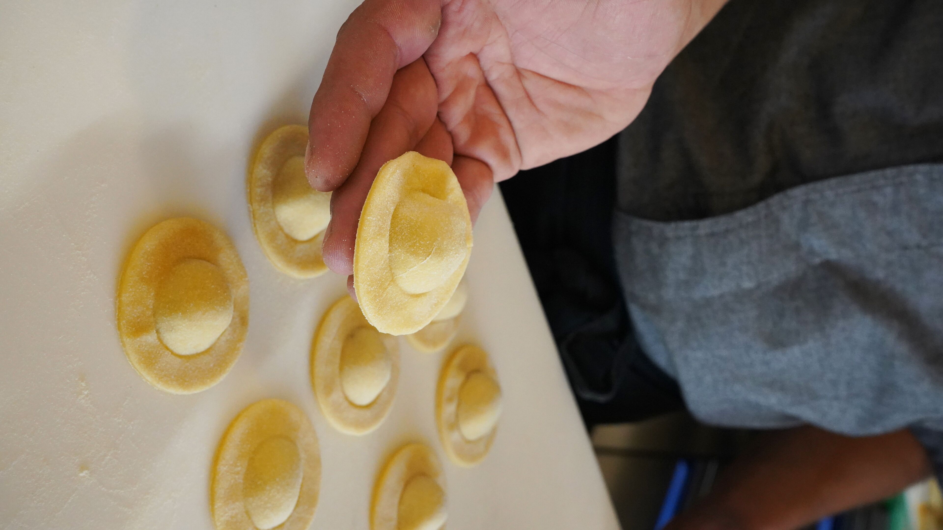 Iemand houdt een ravioli vast op een witte ondergrond, omringd door meerdere andere ravioli.