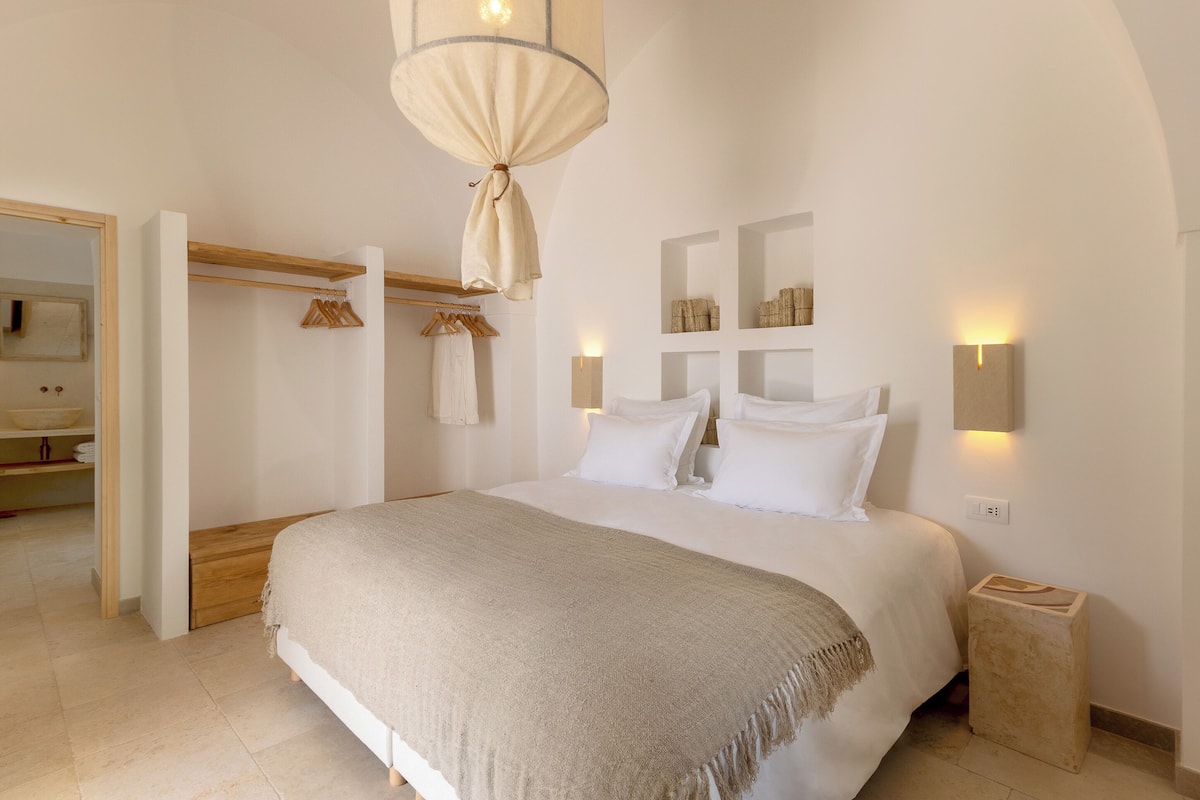A bedroom with a large bed, a hanging light fixture, wall-mounted sconces, and a wooden bench.