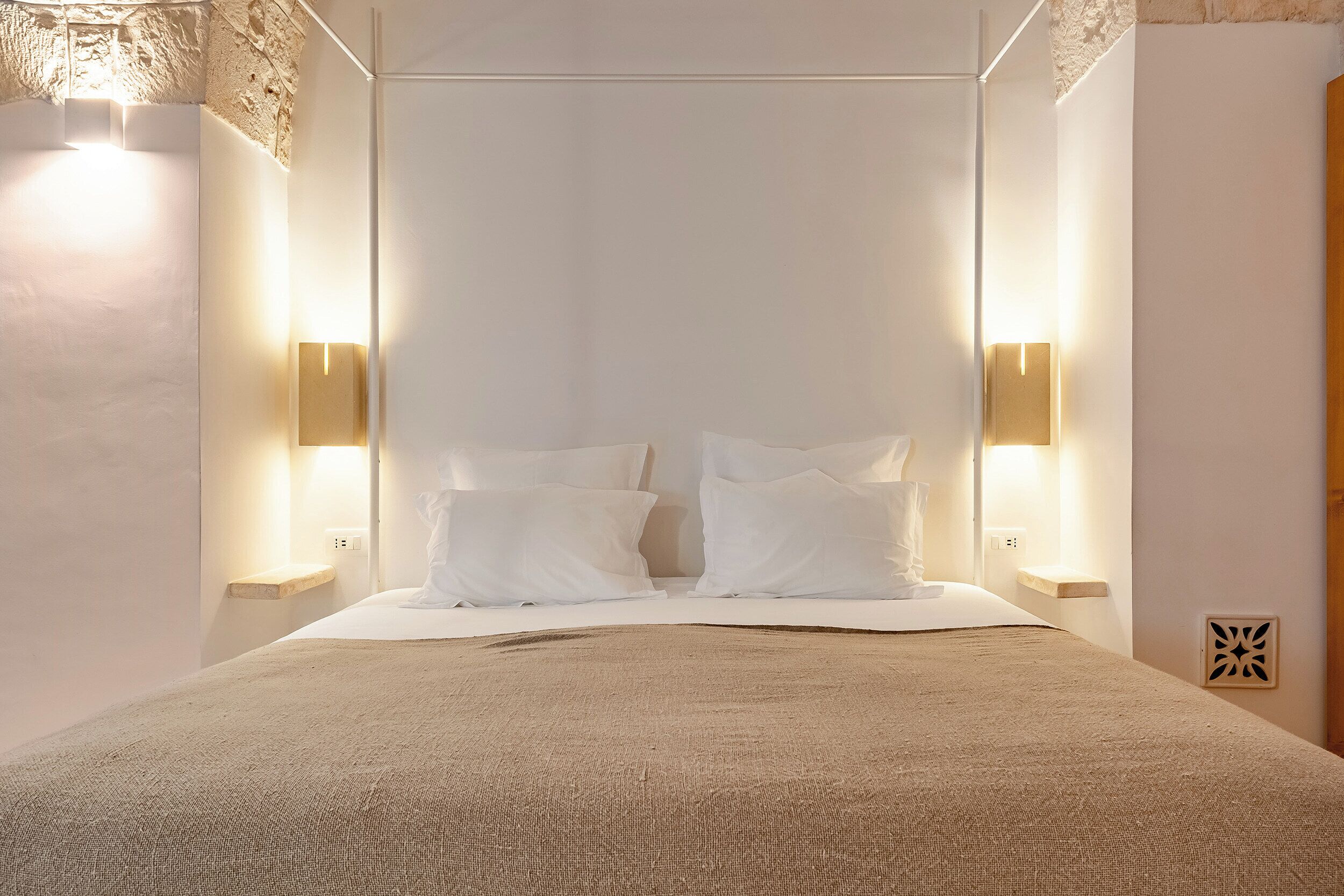 A neatly made bed with white linens, two wall-mounted light fixtures, and a textured wall.