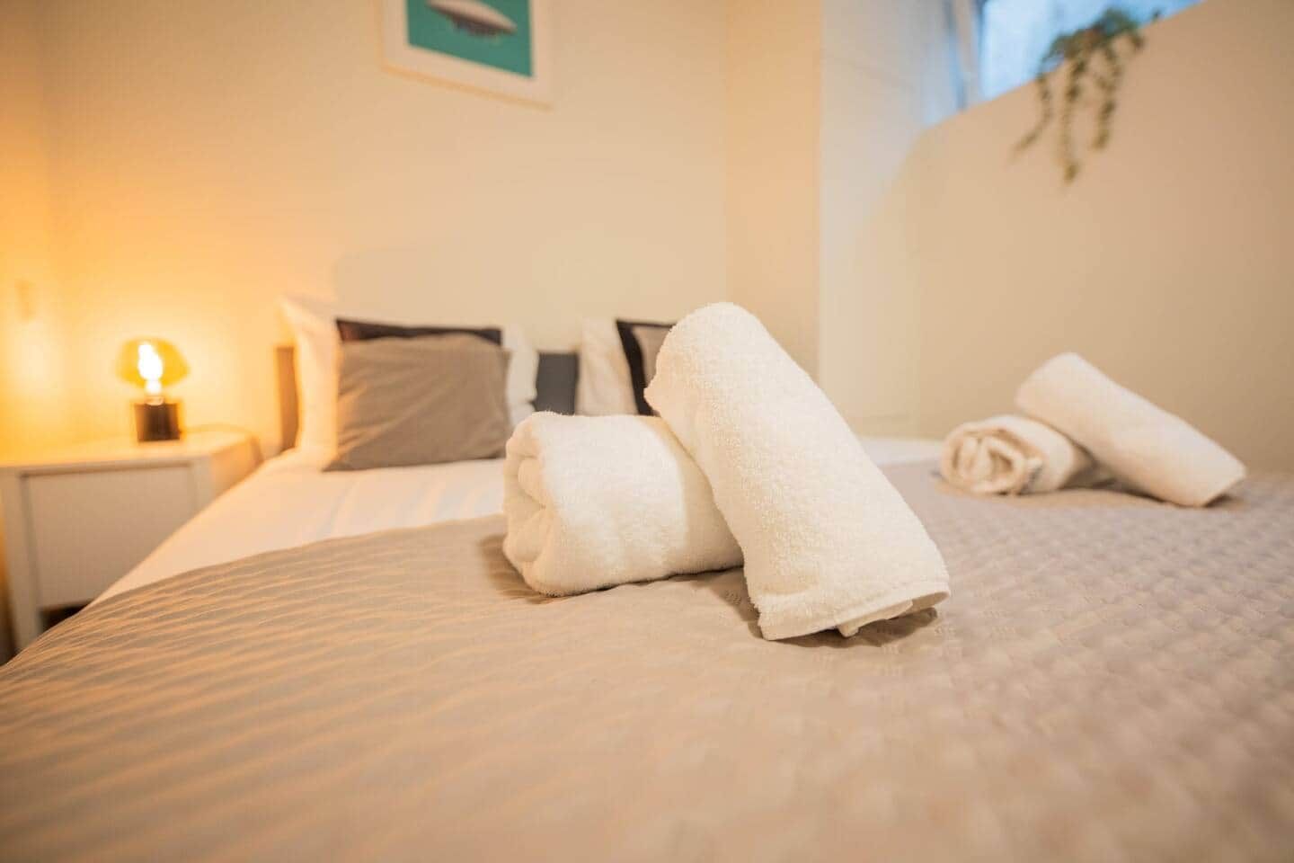 Een net opgemaakt bed met opgerolde handdoeken, een bedlampje en een ingelijste foto aan de muur.