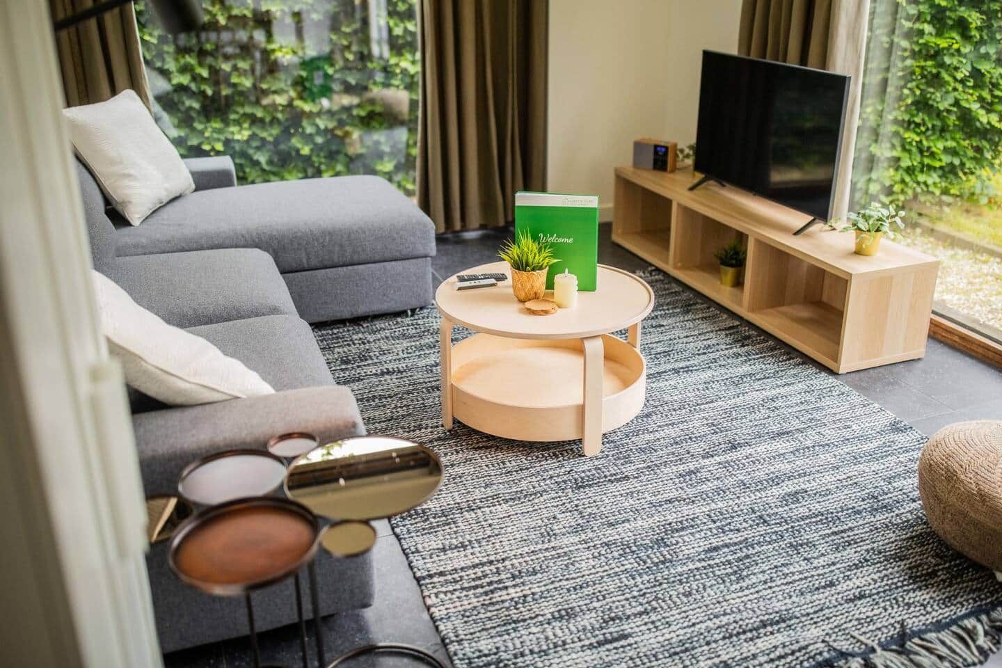 Uma sala de estar moderna com um sofá cinzento, uma mesa de centro redonda, uma TV sobre um suporte de madeira e vista para o verde lá fora.