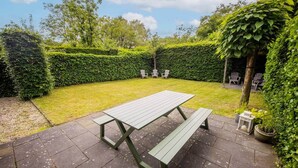 Un espace extérieur aménagé avec une table de pique-nique et des bancs, entouré d’une haie et d’arbres.
