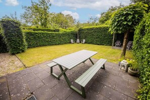 Een terras met een picknicktafel en banken, omgeven door een haag en bomen.