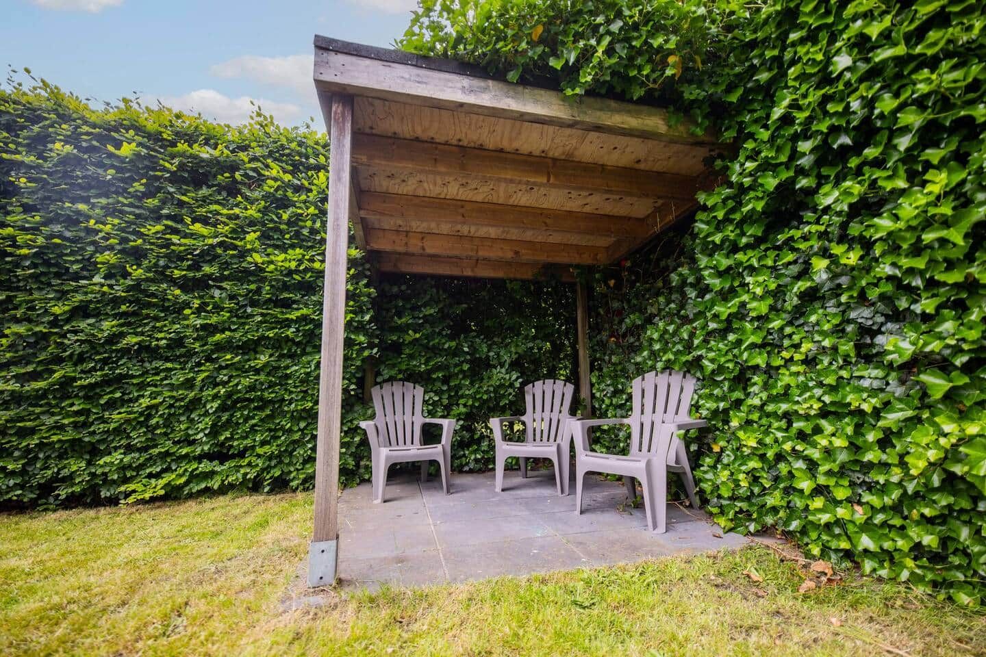 A covered patio with two Adirondack chairs, surrounded by dense green foliage.