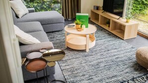 A modern living room with a grey sofa, a round coffee table, a TV on a wooden stand, and a view of greenery outside. - Superb 90m² Apartment with Garden (KS-8-A) (Eindhoven)