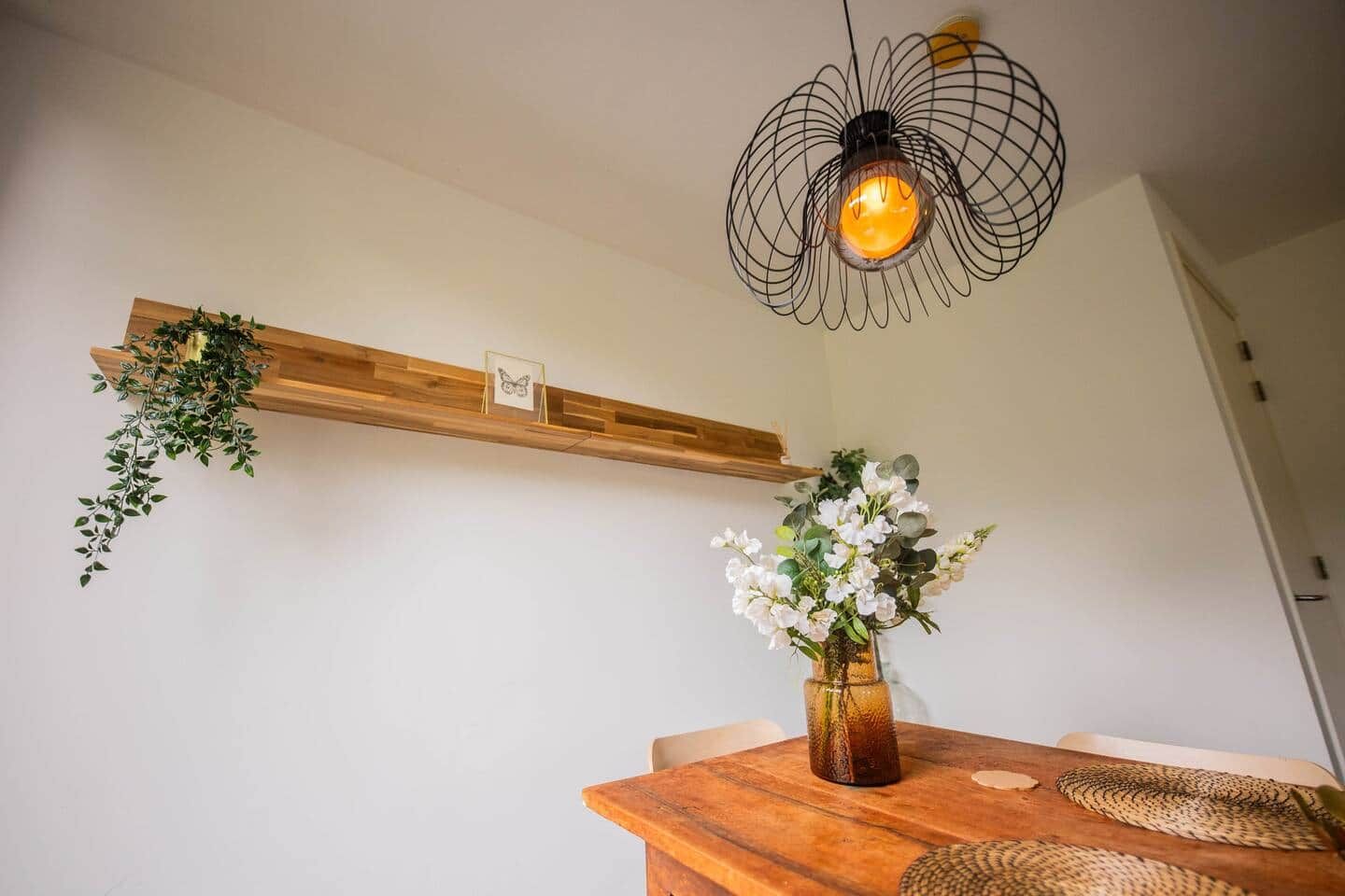 Een houten eettafel met een vaas witte bloemen, een hanglamp en een houten plank met planten.