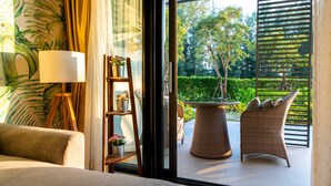 A room with a bed, a lamp, a shelf with plants, and a view of an outdoor patio with wicker furniture and greenery. - Cassia Residences by NLA (Choeng Thale)