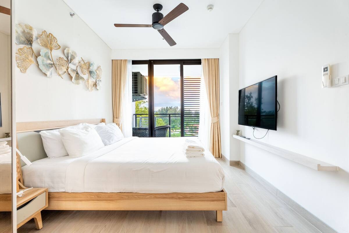 A modern bedroom with a large bed, a ceiling fan, a TV, and a balcony with a view.