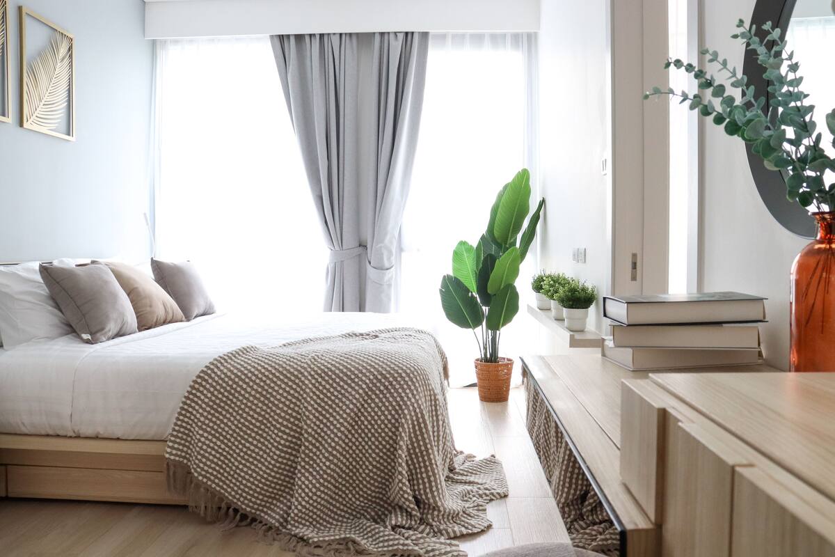 A bedroom with a bed, a large window with curtains, a potted plant, and a bedside table with books.