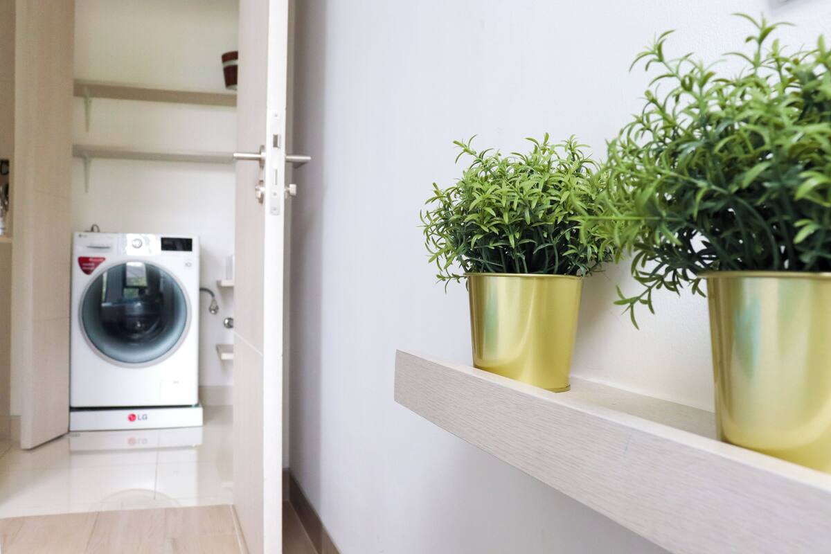 A laundry room with a front-loading washer and dryer, a shelf with two potted plants, and a white wall.