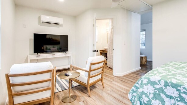 Quarto de hotel com uma cama, televisão, ventoinha no teto e uma pequena mesa com cadeiras.