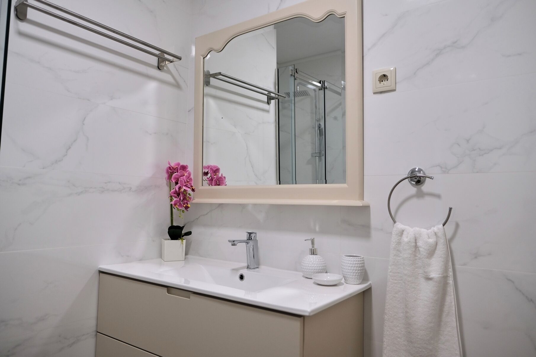 A bathroom with a marble wall, a sink with a mirror, a towel rack, and a shower visible through the glass door.