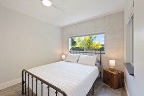 Un dormitorio con cama, dos lámparas de noche, una ventana con vista a follaje e iluminación en el techo.
