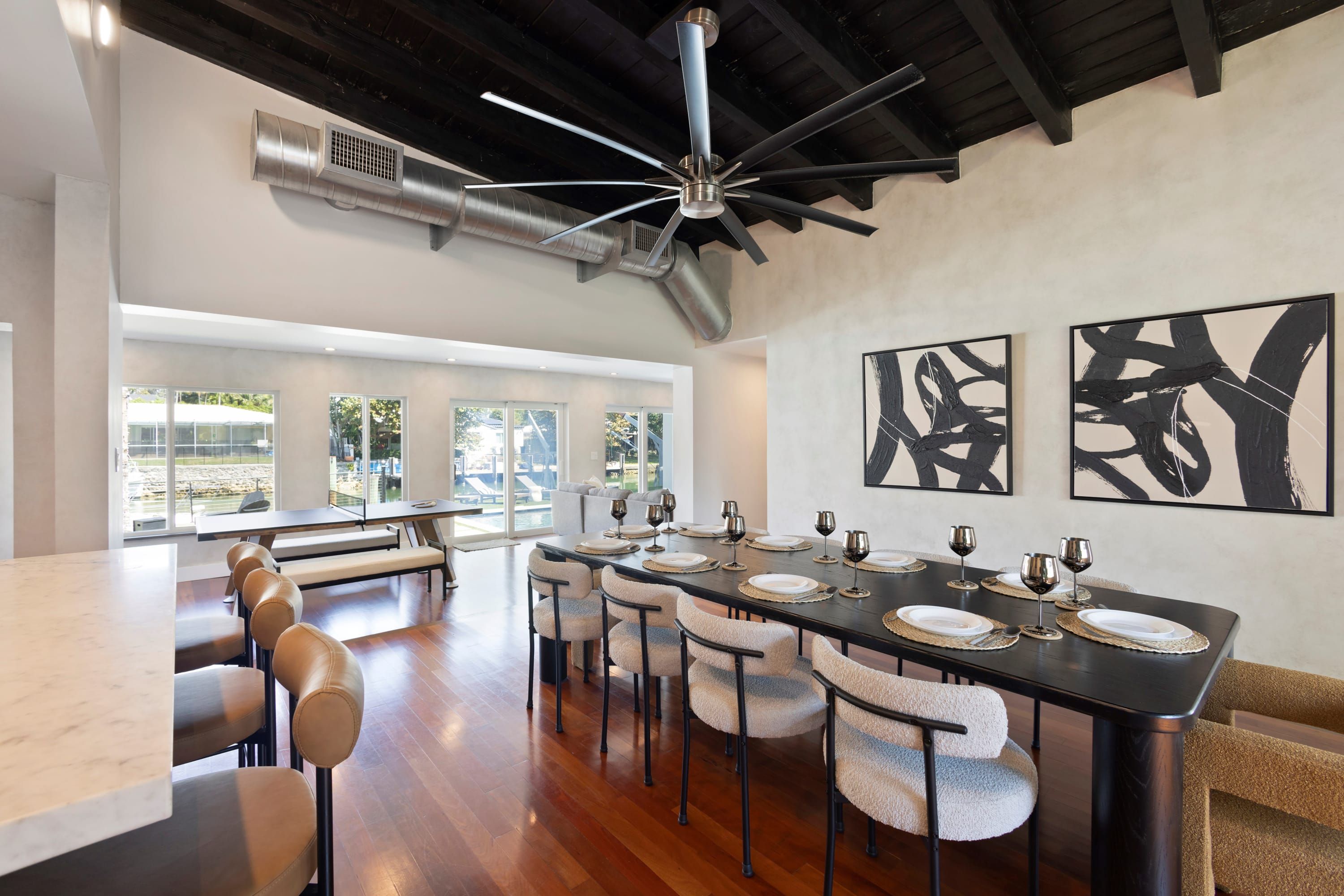 Un coin repas avec une longue table dressée pour six personnes, entourée de chaises rembourrées, un ventilateur de plafond et des œuvres d’art abstraites accrochées aux murs.