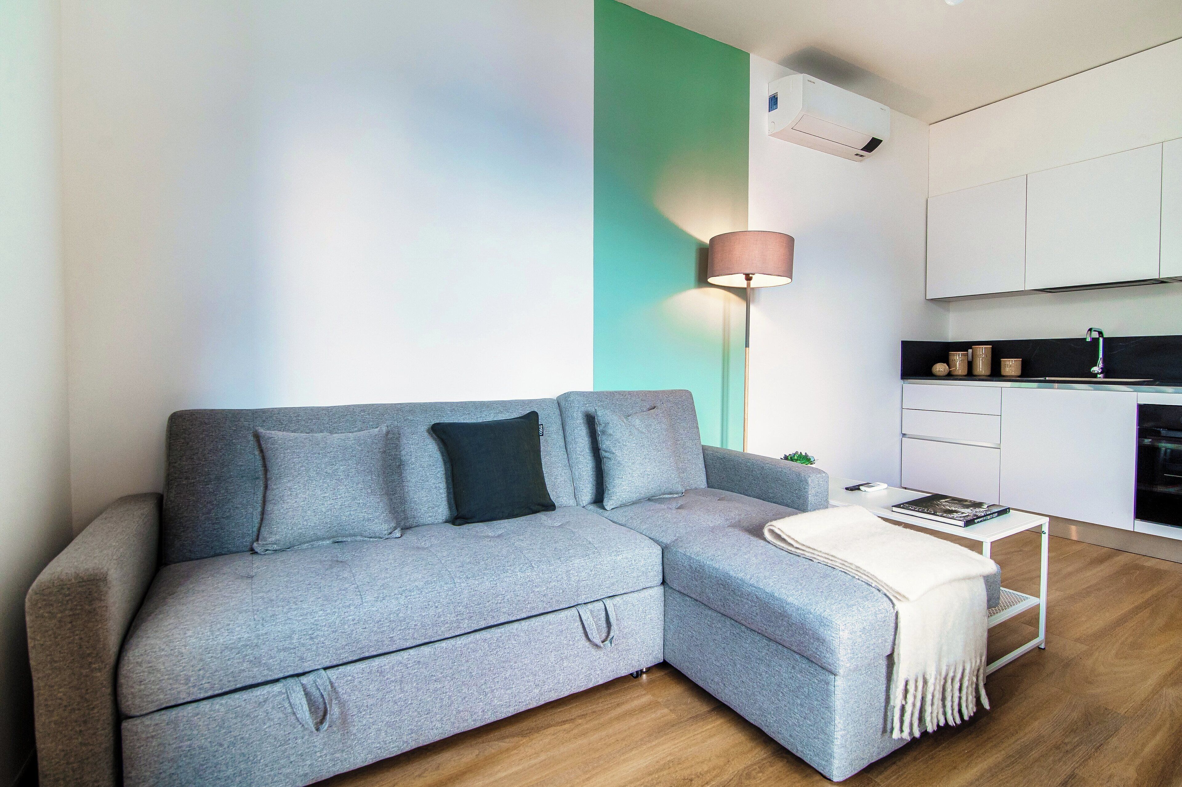 Ein modernes Wohnzimmer mit einem grauen Ecksofa, einem kleinen weißen Beistelltisch und einer Stehlampe.